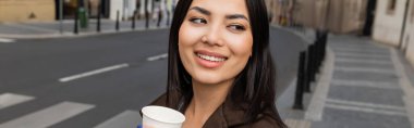 Smiling brunette woman in coat holding paper cup and looking away on blurred urban street, banner  clipart