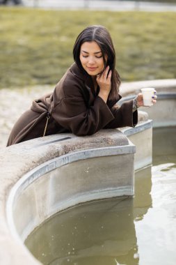 Brunette woman in coat holding paper cup near fountain in Prague  clipart