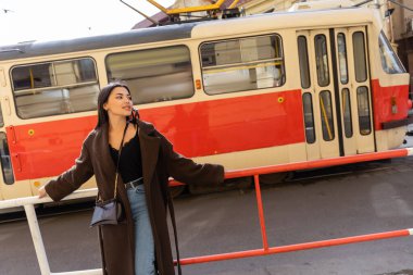 Trendy tourist in coat looking away near tram on urban street in Prague  clipart