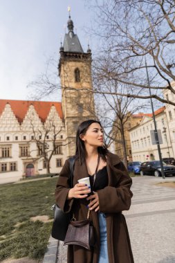 Tourist with paper cup walking on urban street and looking away in Prague  clipart