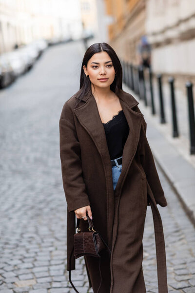 stylish brunette woman in brown coat holding handbag while walking on street in prague