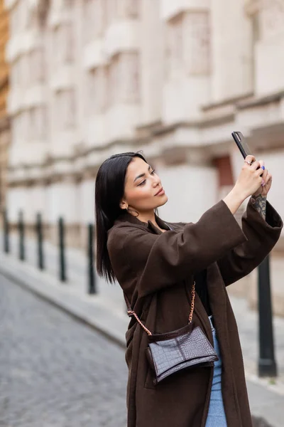 stock image pretty young woman in brown coat taking selfie in prague on blurred background