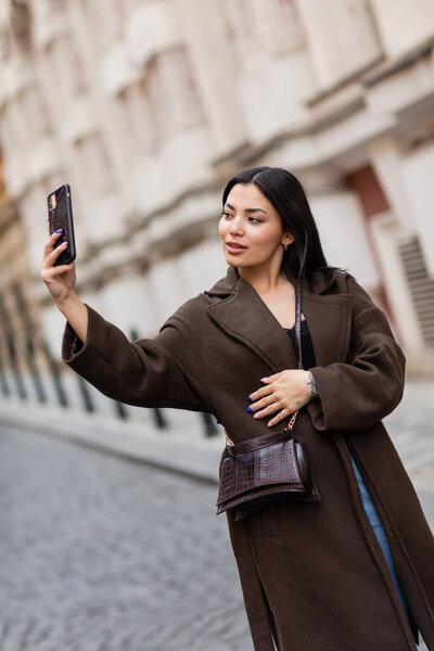 young brunette woman in brown coat taking selfie on blurred street in prague