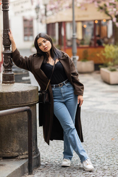 young brunette woman in brown coat and jeans leaning on lamppost on street in prague