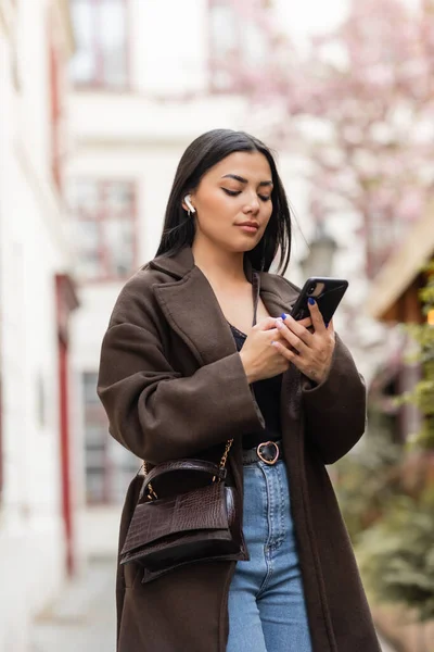 Stock image trendy brunette woman in wireless earphone using cellphone on street in prague