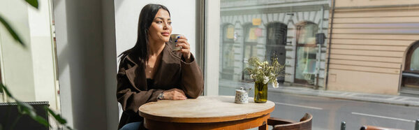brunette woman in coat drinking coffee and looking through window in prague cafe, banner
