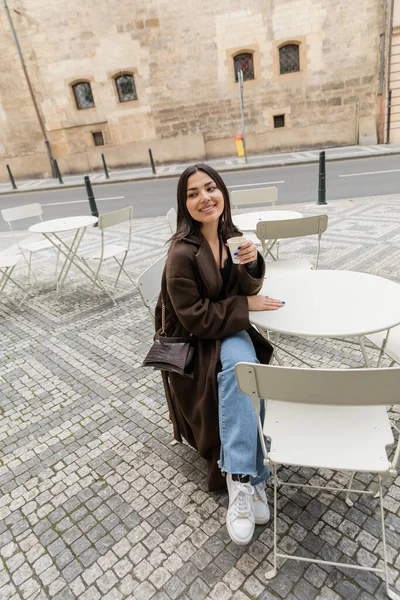 Smiling traveler in coat holding paper cup near table of outdoor cafe in Prague