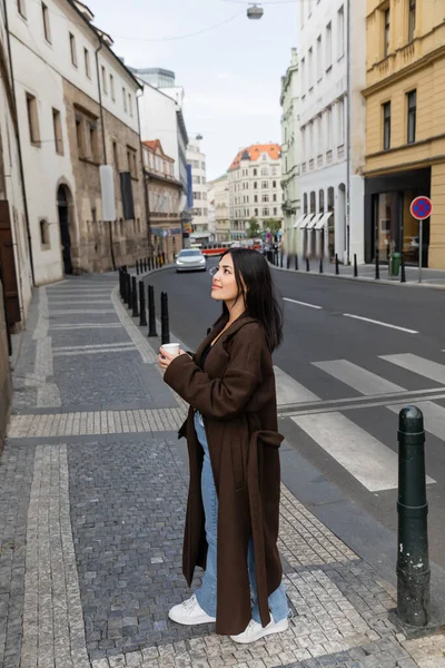 Pretty traveler in coat holding paper cup and looking away on urban street in Prague