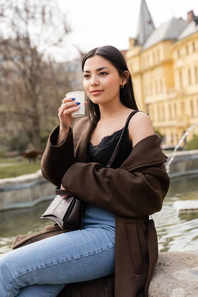 Stylish woman in coat holding paper cup near blurred fountain on street in Prague