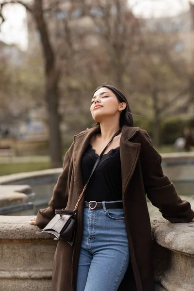 stock image Young woman in coat standing near blurred fountain on street in Prague 