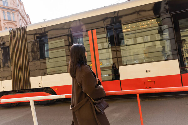 Young brunette woman in coat looking at tram on street in Prague 