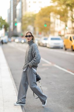 full length of stylish woman in sunglasses and grey outfit holding handbag while walking on street of New York city  clipart