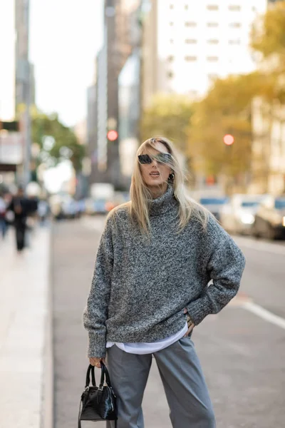 stock image blonde woman in sunglasses and trendy outfit holding handbag while posing with hand on hip on street of New York city 