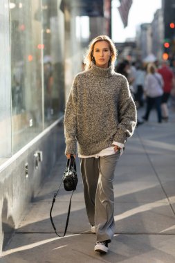 full length of young and stylish woman in grey outfit walking with trendy handbag on street of New York  clipart