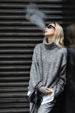 young woman in winter sweater and sunglasses smoking e-cigarette while standing with handbag on urban street  clipart