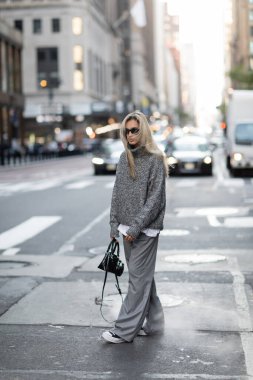 Uzun boylu, örgü süveter ve güneş gözlüklü sarışın bir kadın New York 'ta şehir sokağında son moda bir çantayla yürüyor. 