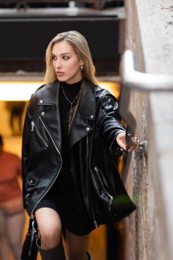 stylish young woman in leather jacket and black dress holding metal handrail near subway entrance in New York  clipart