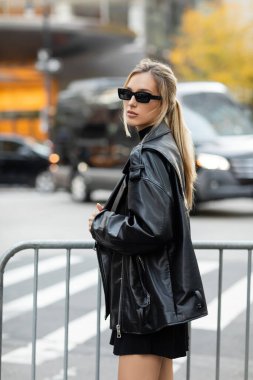 stylish woman in black leather jacket and sunglasses standing near metal barrier in New York  clipart