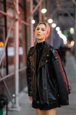 blonde woman in black leather jacket standing on street in New York city at evening time clipart