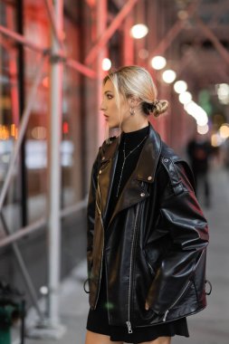 side view of blonde woman in black leather jacket standing on street in New York city at evening time clipart