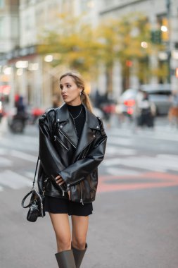 stylish woman in leather jacket and dress walking near blurred cars on street in New York city  clipart