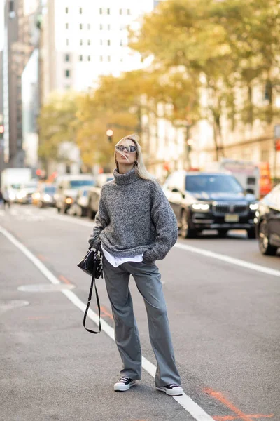 stock image full length of stylish woman in sunglasses and grey outfit holding black handbag and standing on street of New York city 