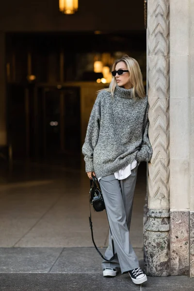 full length of blonde woman in sunglasses and grey outfit holding handbag while standing near building in New York