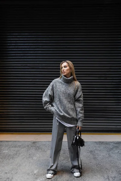 stock image full length of young woman in winter outfit standing with hand in pocket and holding handbag outdoors 