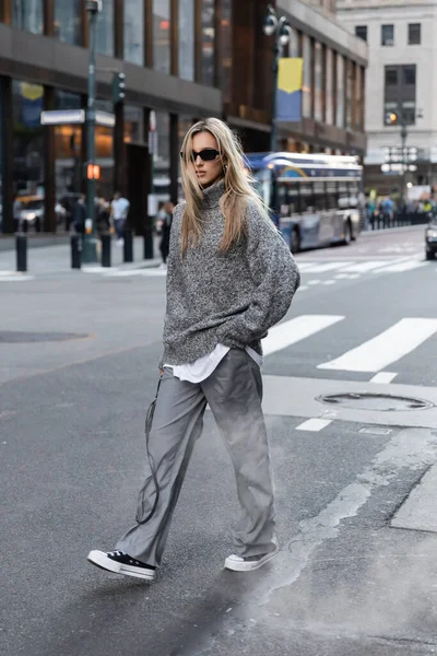 stock image full length of blonde woman in stylish knitted sweater and sunglasses walking on urban street in New York 