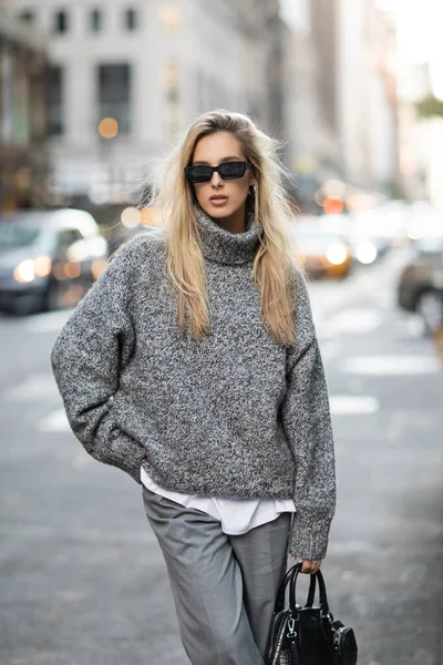 stock image blonde woman in knitted sweater and sunglasses standing with trendy handbag on blurred street in New York 