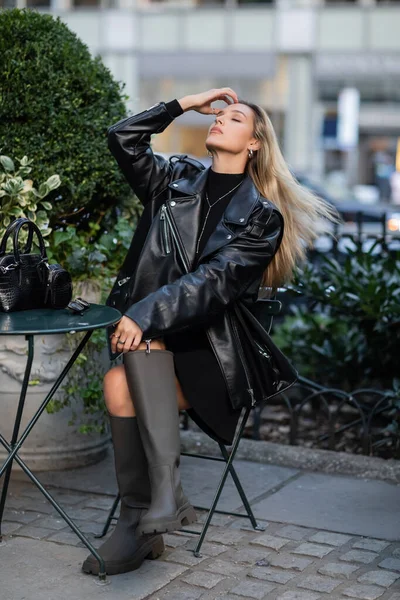 full length of stylish woman in leather jacket and boots sitting in outdoor cafe on street of New York