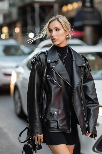 stylish woman in leather jacket and black dress walking near blurred cars on street in New York