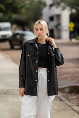 young blonde woman in stylish outfit looking at camera while standing on street in Miami  clipart