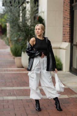 full length of young woman in leather shirt jacket and cargo pants posing with handbag on street in Miami  clipart