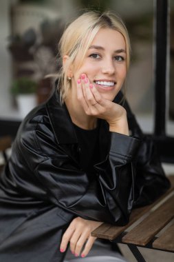 cheerful young woman in leather shirt jacket sitting in outdoor cafe on street in Miami clipart