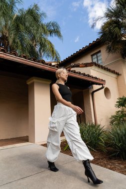 full length of young woman in trendy clothes walking in trendy boots near modern house in Miami  clipart