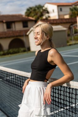 Kolsuz tişörtlü, beyaz kargo pantolonlu, tenis kortunda ağlara yaslanmış neşeli genç bir kadın.