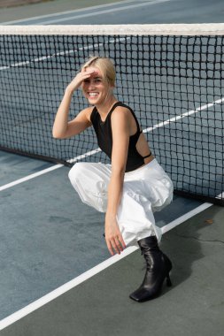 Uzun kollu tişörtlü ve beyaz kargo pantolonlu mutlu bir kadın tenis kortunda ağın yanında oturuyor.