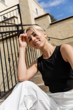 cheerful woman in tank top and white cargo pants smiling near building in Miami  clipart