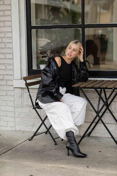 full length of young woman in leather shirt jacket and cargo pants sitting in outdoor cafe on street in Miami 