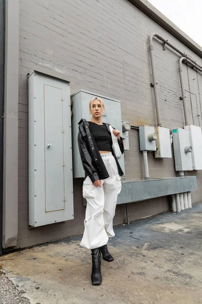 stock image full length of young blonde woman in leather shirt jacket and cargo pants standing with handbag on industrial street in Miami 