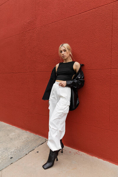 full length of blonde woman in cargo pants and leather shirt jacket standing near red wall in Miami 