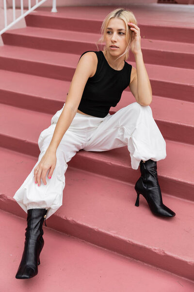 full length of blonde young woman in black tank top and cargo pants sitting on pink stairs in Miami 