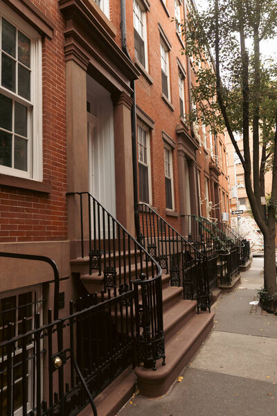 кирпичное здание с белыми окнами и входами с лестницами и металлическими заборами возле дерева на городской улице в Нью-Йорке