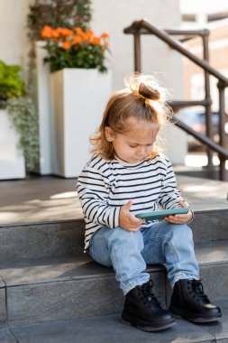 Evin yakınındaki merdivenlerde otururken akıllı telefon kullanan günlük kıyafetli küçük bir kız. 