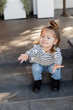 Evin yakınındaki merdivenlerde otururken dikkat çekmek ve el kol hareketi yapmak isteyen günlük kıyafetli bir kız. 