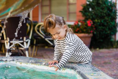 toddler kid in striped long sleeve shirt wetting hands in water of fountain in Miami  clipart