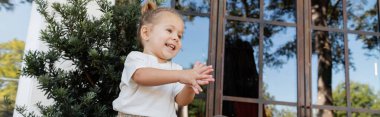 positive toddler girl in white t-shirt clapping hands near outdoor cafe in Miami, banner  clipart