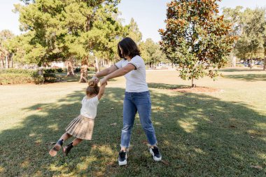 full length of brunette woman in jeans playing with toddler daughter in park of Miami  clipart