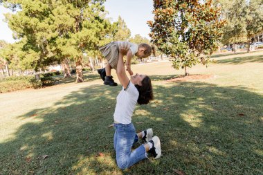 full length of cheerful mother in jeans lifting toddler daughter while playing in park of Miami  clipart